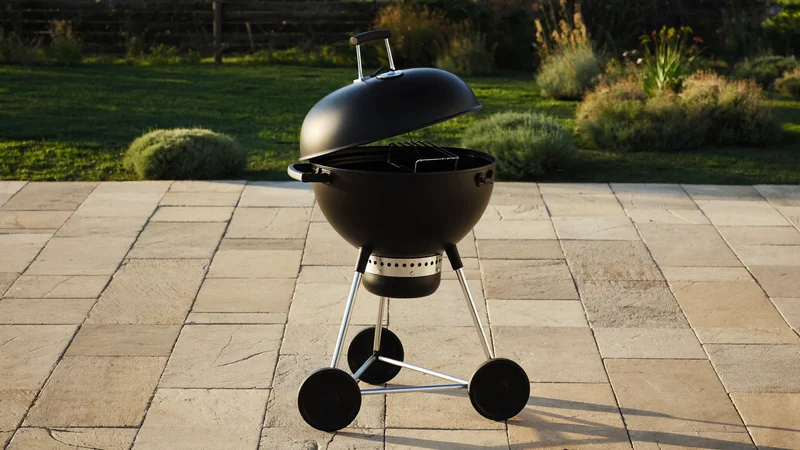 Barbecue kettle noir mat sur une terrasse dallée, couvercle légèrement ouvert