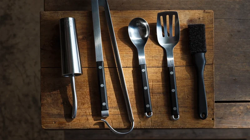 Pinces, spatule, fourchette et pinceau inox alignés sur une planche en chêne